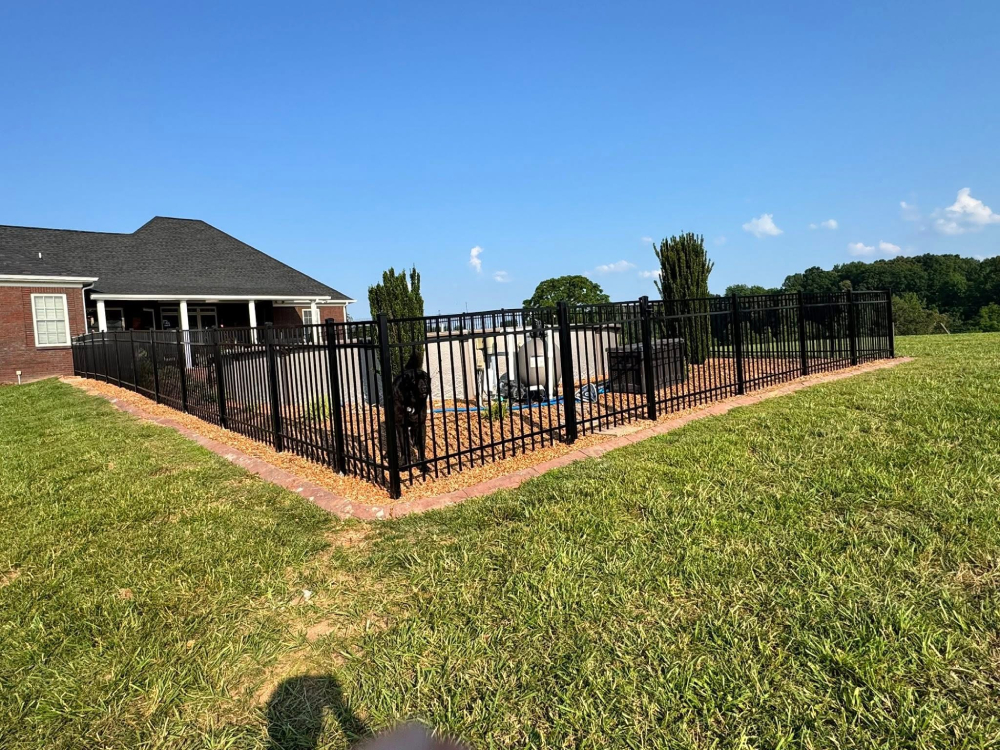 A black metal fence surrounds a backyard pool next to a brick house, with green grass and trees in the background under a clear blue sky. | Supremo Exteriors