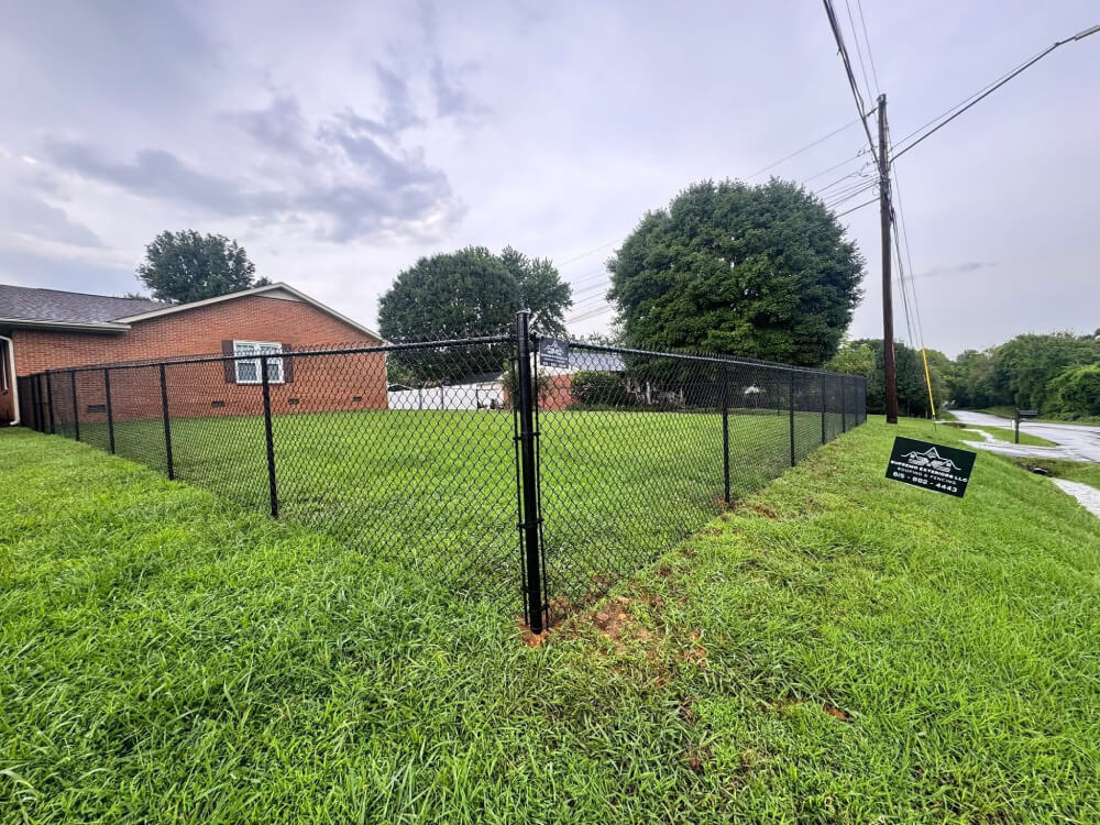 A neatly mowed backyard enclosed by a black chain-link fence next to a brick house. There are large green trees and a sign attached to the fence near a street with tall utility poles. The sky is overcast. | Supremo Exteriors