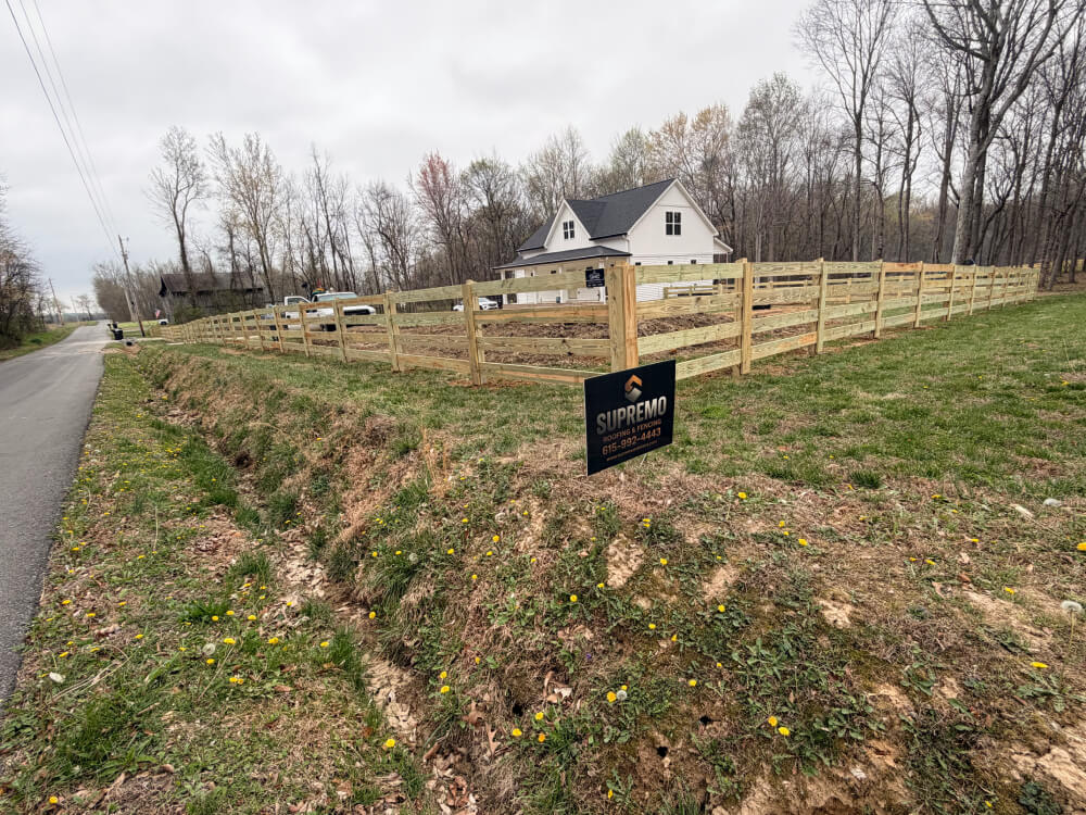 A fenced yard with a white house in the background, surrounded by trees. A black sign in the foreground reads SUPREMO FENCING. The scene is overcast with patches of grass and dandelions. | Supremo Exteriors