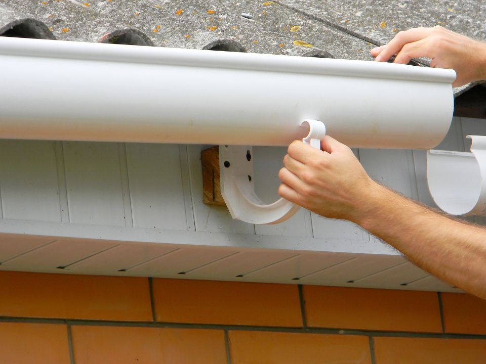 A person’s hands attach a white plastic gutter to a roof edge using a bracket. The roof has gray corrugated tiles, and the wall below is made of orange bricks. | Supremo Exteriors