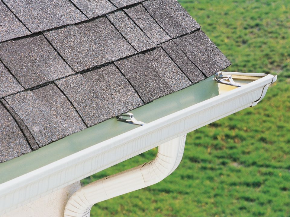 Close-up of a house roof with gray asphalt shingles and a white rain gutter system, including gutter brackets and a downspout, set against a background of green grass. | Supremo Exteriors
