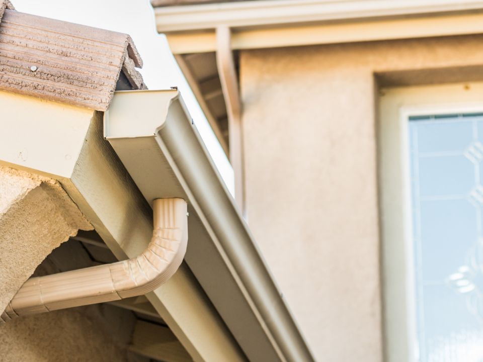 Close-up of a house roof edge showing beige rain gutters and downspout attached to the side, with part of a textured wall, window, and roof tiles visible in the background. | Supremo Exteriors