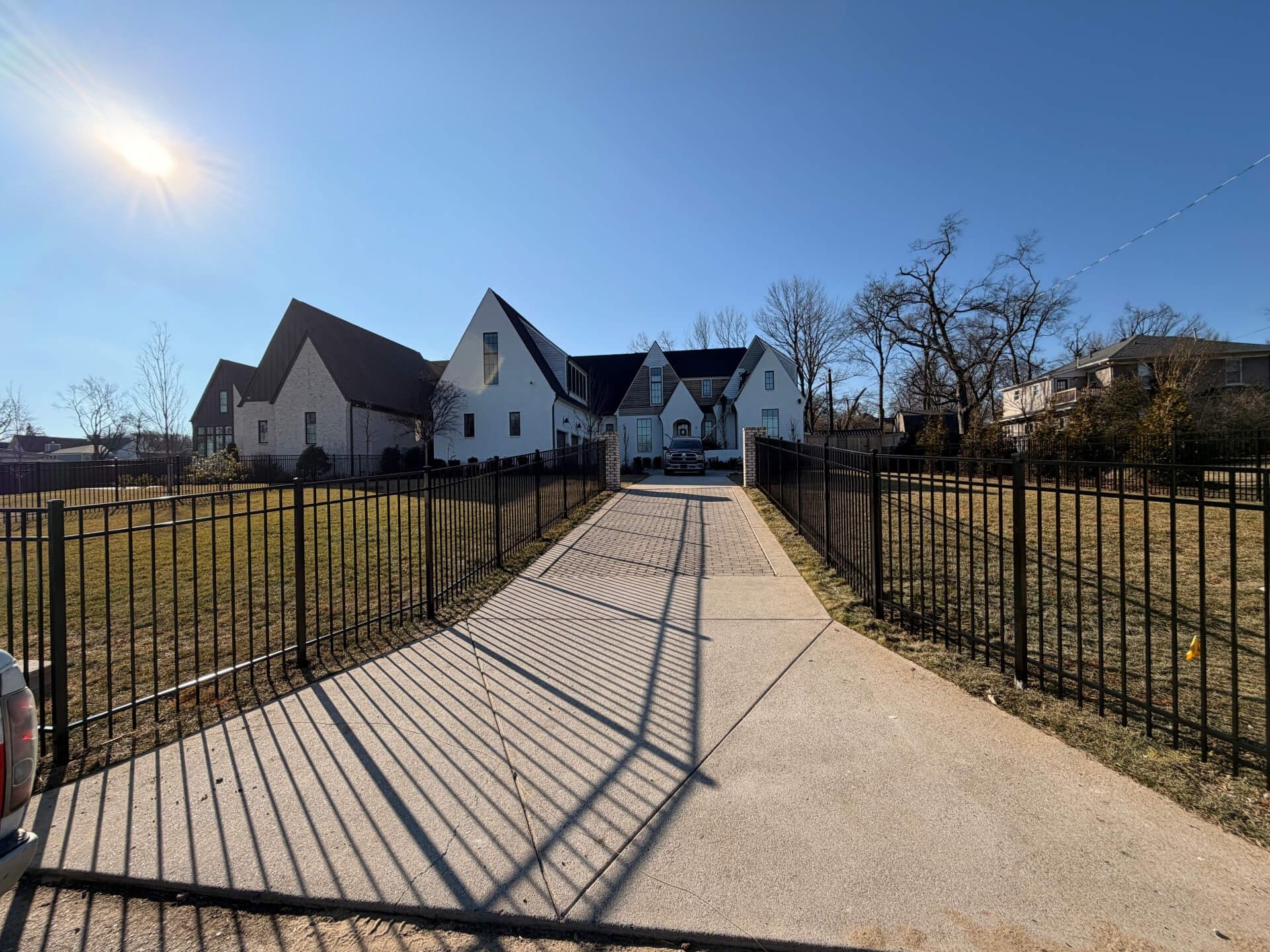 A wide concrete driveway leads to a large white house with dark roofs, fenced on both sides. The sun is shining brightly in a clear blue sky, casting long shadows from the fence onto the driveway. | Supremo Exteriors