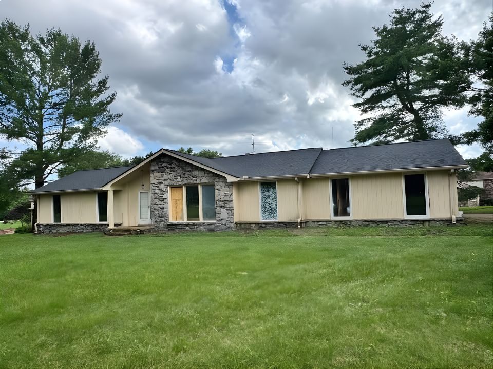 A single-story house with light-colored siding, a stone accent facade, and several large windows. One window is boarded up, and another is covered with a blue tarp. The house sits on a grassy lawn with trees in the background. | Supremo Exteriors