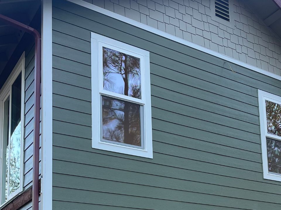 A green house exterior with white trim, featuring a double-hung window reflecting trees. The siding is horizontal on the lower section and shingled on the upper section near the roof. | Supremo Exteriors