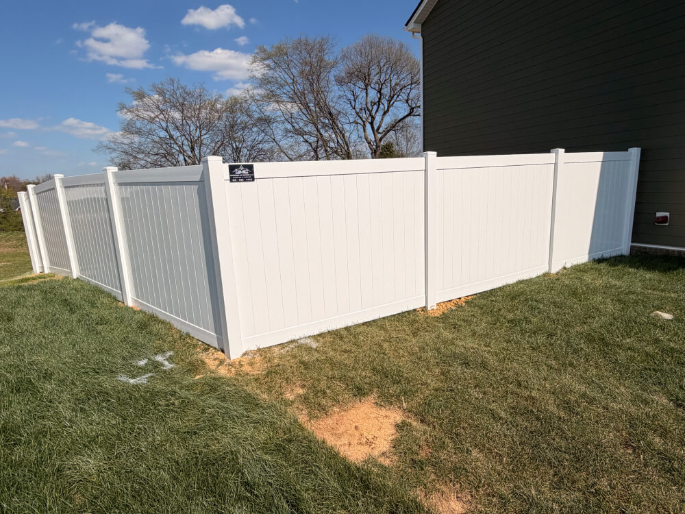 A white vinyl privacy fence encloses a grassy backyard area next to a house, with patches of dirt near the fence and trees visible in the background under a blue sky. | Supremo Exteriors