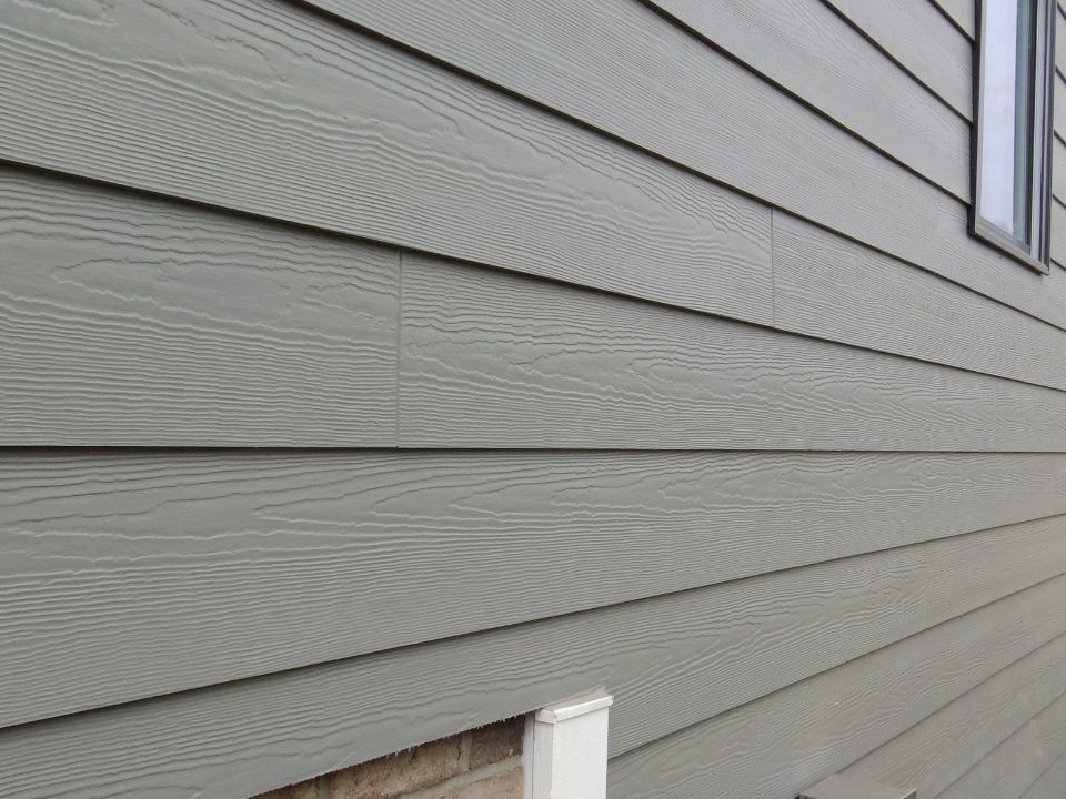Close-up of gray horizontal siding on a house exterior, showing wood grain texture, with part of a window and a small white vent or utility box near the bottom. | Supremo Exteriors