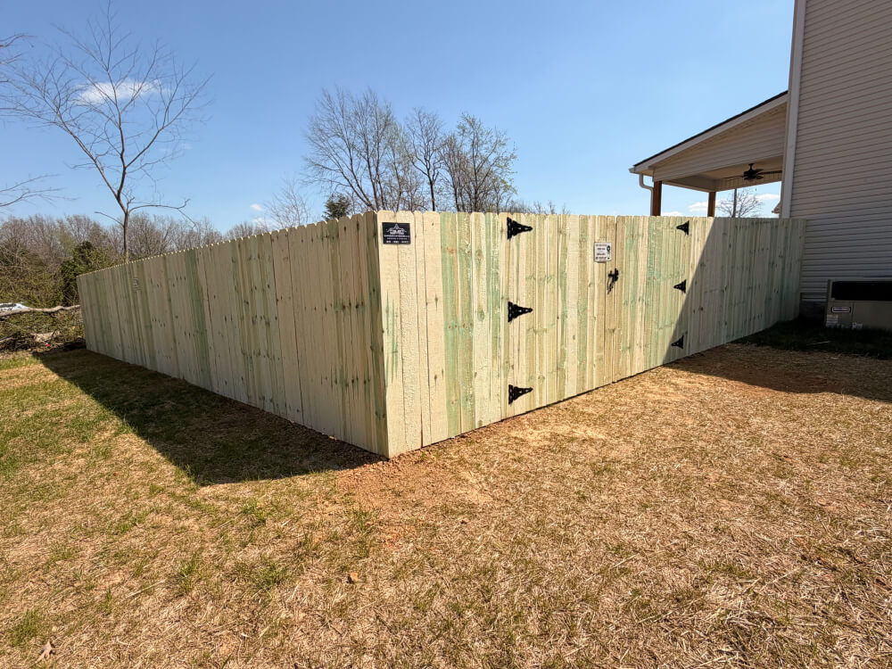A newly built wooden privacy fence surrounds a backyard next to a house. The fence has clean, light-colored wood panels, metal hinges, and latches on a gate. Bare trees and a patchy yard are visible under a clear sky. | Supremo Exteriors