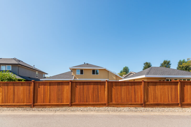 A row of suburban houses with light-colored exteriors and dark roofs stands behind a tall, wooden privacy fence on a clear, sunny day. | Supremo Exteriors