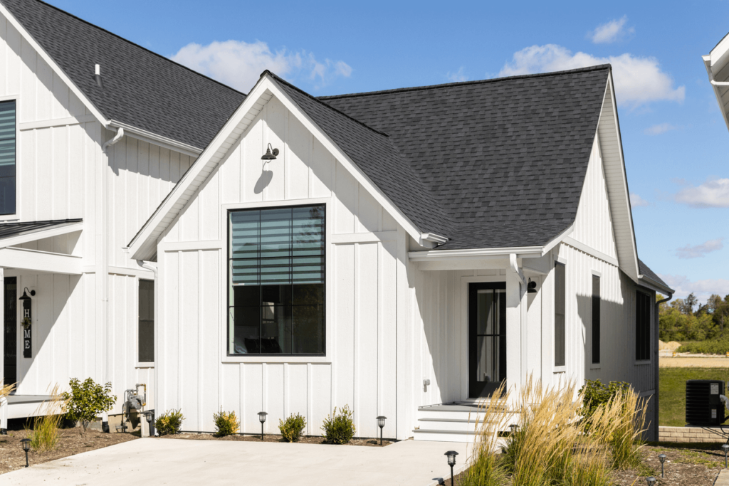 Modern white house with black roof and large front window, featuring simple landscaping, small bushes, ornamental grasses, and a concrete driveway under a clear blue sky. | Supremo Exteriors
