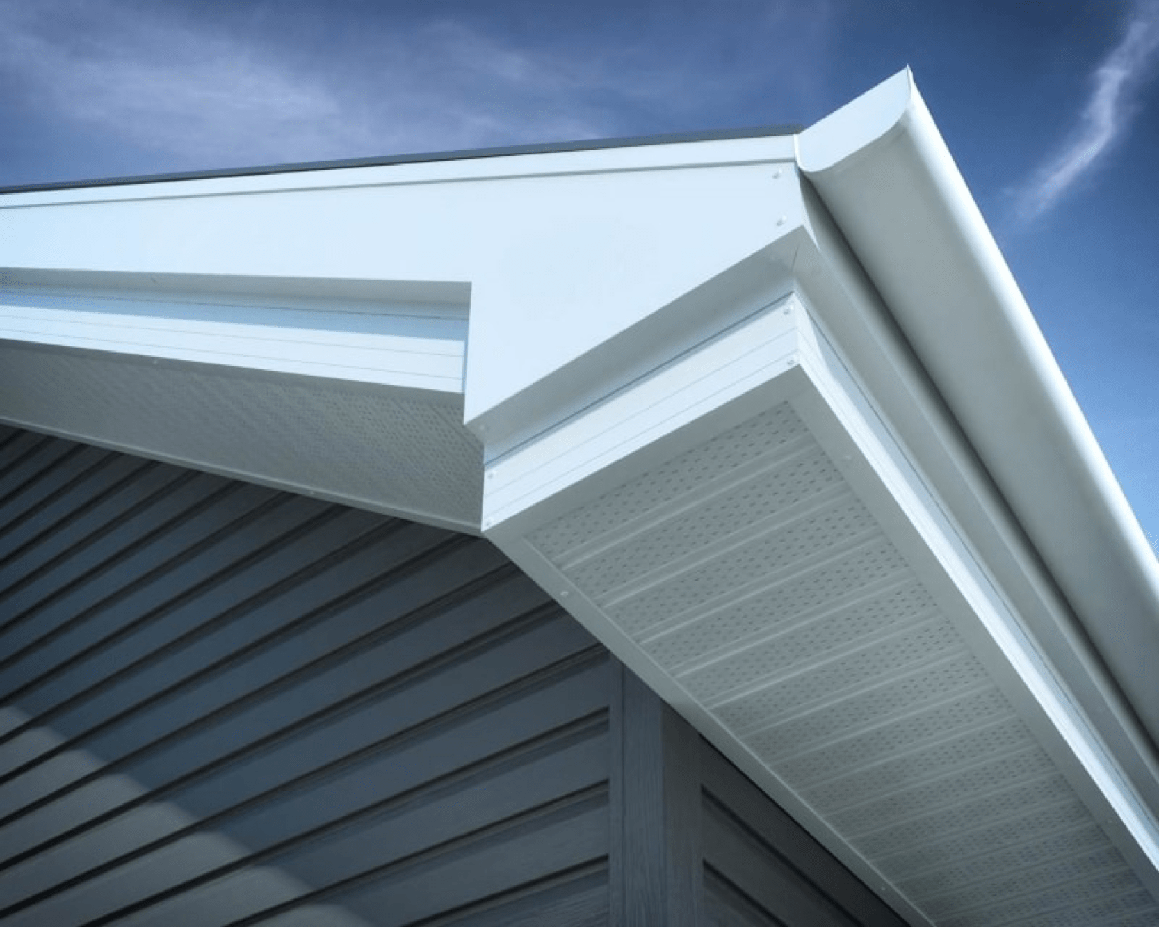 Close-up view of a house’s roof corner, showing white soffit and fascia with gray vinyl siding against a blue sky background. | Supremo Exteriors