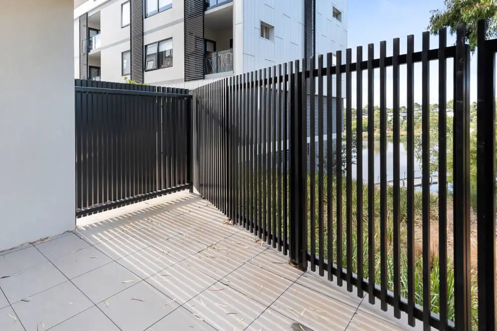 A modern outdoor patio with light-colored floor tiles, surrounded by a black metal fence. An apartment building and greenery are visible in the background. Sunlight casts shadows from the fence onto the tiled floor. | Supremo Exteriors