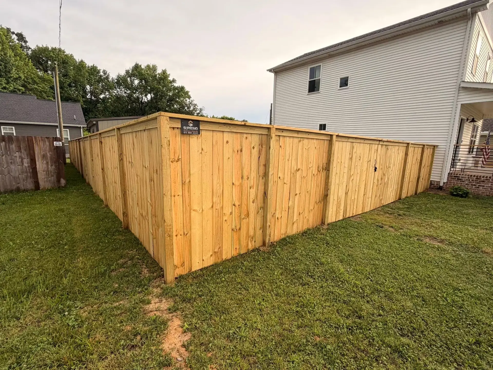 A tall wooden privacy fence encloses a grassy backyard beside a white two-story house, with neighboring houses and trees visible in the background under a cloudy sky. | Supremo Exteriors