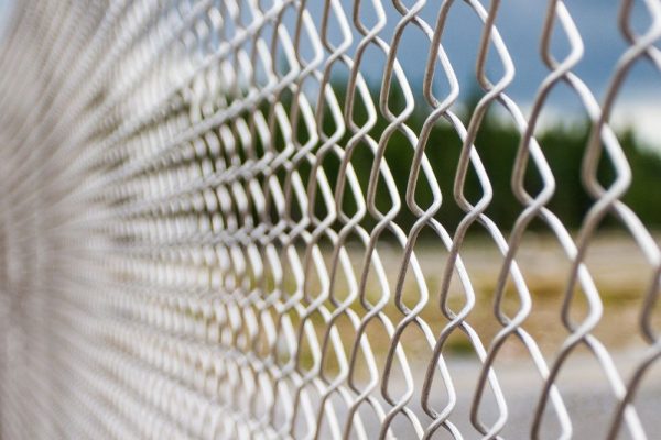 Close-up view of a metal chain-link fence, with a blurred outdoor background of trees and cloudy sky. The fence pattern creates a repeating geometric design. | Supremo Exteriors