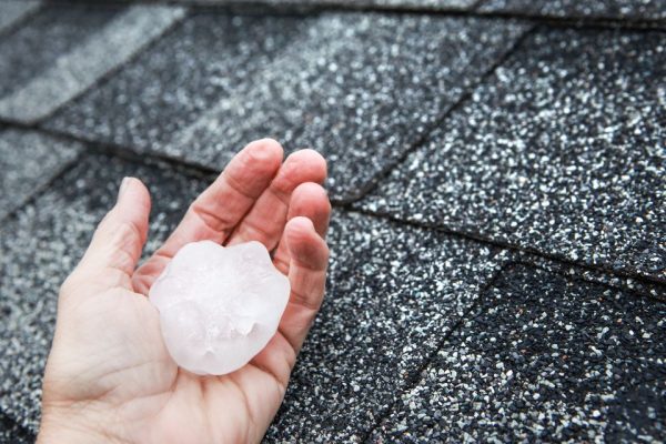 A hand holding a large piece of hail with grey asphalt roof shingles in the background. | Supremo Exteriors