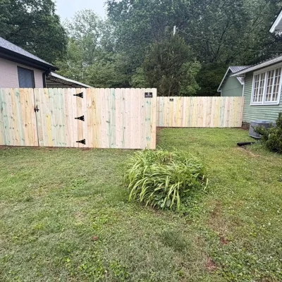 A newly built light wood fence with black metal hinges runs between two houses, enclosing a grassy yard with green plants and trees in the background. | Supremo Exteriors