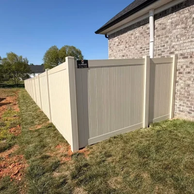 A beige vinyl privacy fence encloses the side yard of a house with light brick siding, set on green grass under a clear blue sky. | Supremo Exteriors