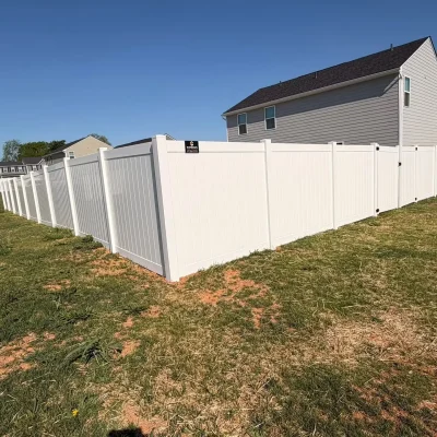 A white vinyl privacy fence encloses the backyard of a light gray house on a sunny day, with green grass and patches of bare soil in the yard. | Supremo Exteriors