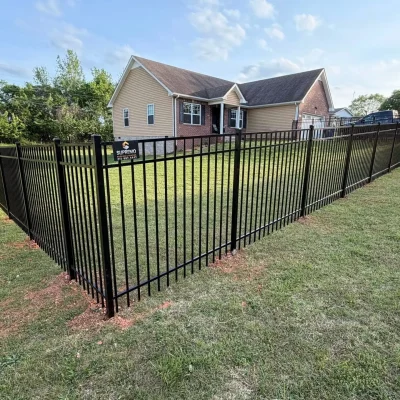 A black metal fence encloses a grassy yard around a tan brick house with white trim on a sunny day. The fence creates a sharp corner in the foreground. Trees and other houses are visible in the background. | Supremo Exteriors