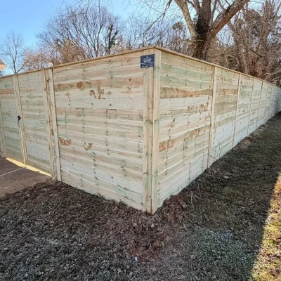 A tall wooden privacy fence with a gate encloses a yard, casting shadows on the ground. Trees and grass surround the fence, and part of a black building is visible on the left. | Supremo Exteriors