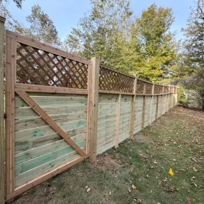 A tall wooden privacy fence with a gate and lattice panels on top runs alongside a grassy yard scattered with fallen leaves. Trees with green and yellow foliage are visible in the background. | Supremo Exteriors