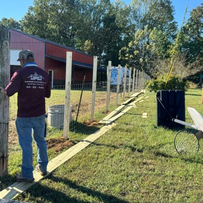 A person installs wooden fence posts along a grassy area near a red barn, with tools and supplies scattered nearby under a clear, sunny sky. | Supremo Exteriors