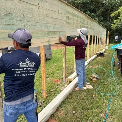 Two workers use power tools to build a wooden fence outdoors near a grassy area. Construction equipment and a truck are visible nearby. | Supremo Exteriors