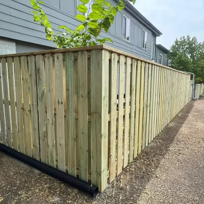 A tall, wooden privacy fence runs alongside a gray house, with a black drainage pipe at the base and a tree growing behind the fence. The ground is a combination of gravel and soil. | Supremo Exteriors