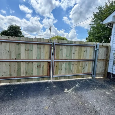 A wooden double gate with metal frame stands on a blacktop driveway beside a white house, under a partly cloudy sky. | Supremo Exteriors