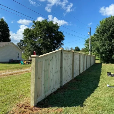 A wooden privacy fence stands on a grassy lawn beside a house, with a blue sky and scattered clouds overhead. Trees and power lines are visible in the background. | Supremo Exteriors