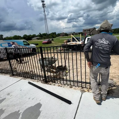 A worker in a gray shirt and hat stands by a black metal fence under construction near a sidewalk. Trucks, tools, and construction materials are visible, with cloudy skies overhead and houses in the background. | Supremo Exteriors