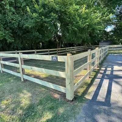 A fenced rectangular area with light wood posts and wire mesh, located beside a paved parking lot with a car, bordered by grass and dense green trees in the background. | Supremo Exteriors