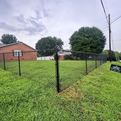 A chain-link fence encloses a grassy yard beside a brick house. Trees line the back of the yard, and a small sign is posted near the road. Power lines run above the yard along the street. | Supremo Exteriors