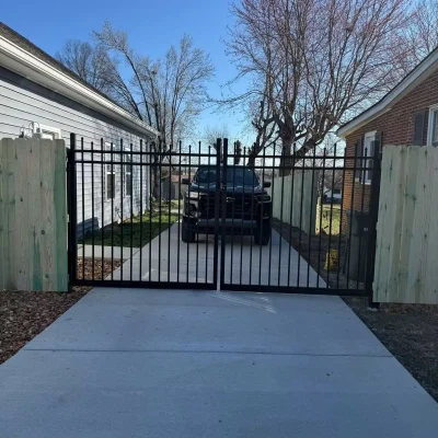 A black metal gate blocks a driveway between two houses, with a black vehicle parked behind the gate. Leafless trees and a clear blue sky are visible in the background. | Supremo Exteriors