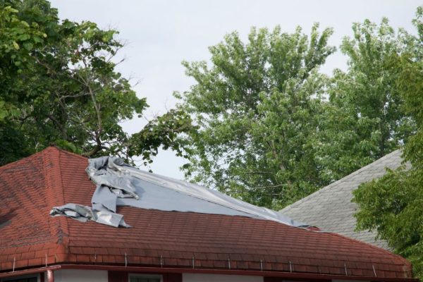 A house with a red roof has part of its roof covered with a tarp, likely for protection or after damage. Green trees surround the house, and another roof is visible in the background. | Supremo Exteriors